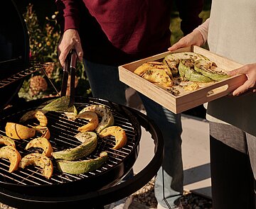 Hände von Menschen, die Gemüse grillen und mit einer Zange vom Grill auf ein hölzernes Tablett legen