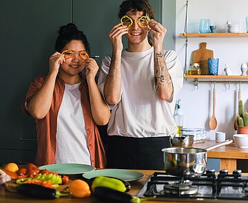 Zwei Menschen nebeneinander in einer Küche, die sich lächelnd Paprika-Stückchen vor die Augen halten