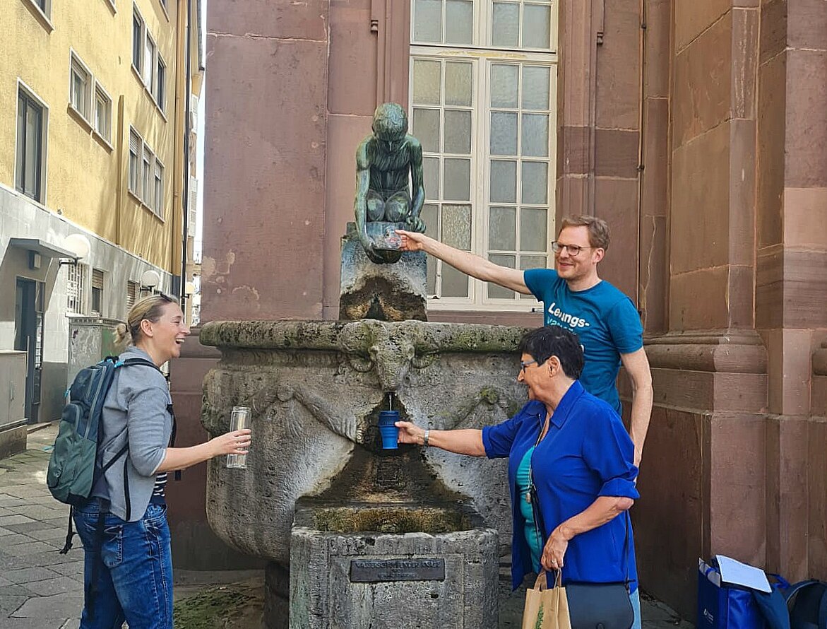 Drei Personen füllen Gläser und Flaschen an Brunnen auf