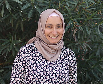 Porträt von Nurdagül Aras-Altas für die KA°-Community - eine kopftuchtragende lächelnde Frau in einem Kleid vor einem Oleander
