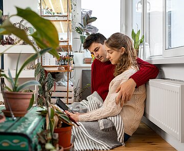 Mann und Frau sitzen Arm in Arm an Heizkörper und schauen gemeinsam auf Tablet