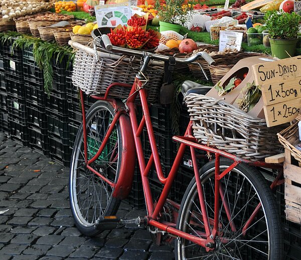 Ein Fahrrad lehnt an einen Marktstand. Die Fahrradkörbe (vorne und hinten) sind reich gefüllt mit Obst und Gemüse
