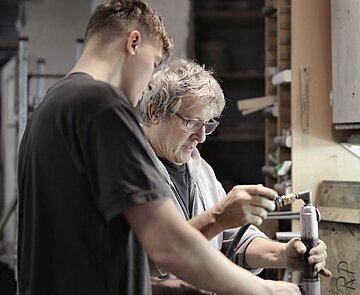 Eine jüngere, männlich gelesene Person hilft einer älteren, männlich gelesenen Person bei einer handwerklichen Aufgabe.