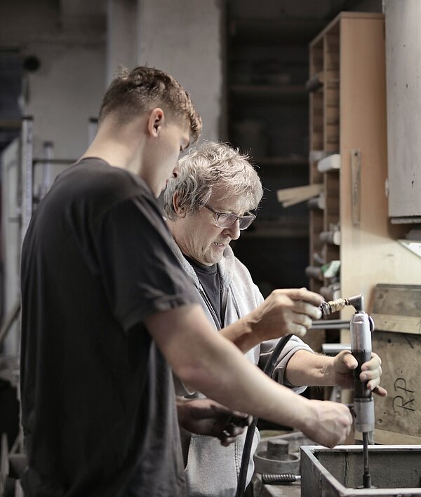 Eine jüngere, männlich gelesene Person hilft einer älteren, männlich gelesenen Person bei einer handwerklichen Aufgabe.