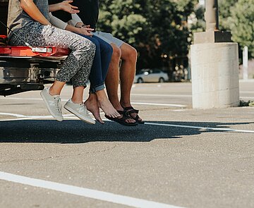 drei Personen, die auf einem Parkplatz Ladefläche eines Autos sitzen - man sieht nur die Beine hinunter baumeln