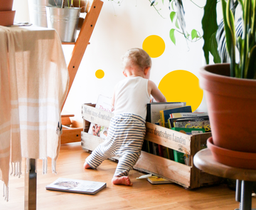 Tatenbank wohnen - Kind in einem Wohnzimmer, Bücher aus einer Kiste räumend, gelbe Sphären an der Wand