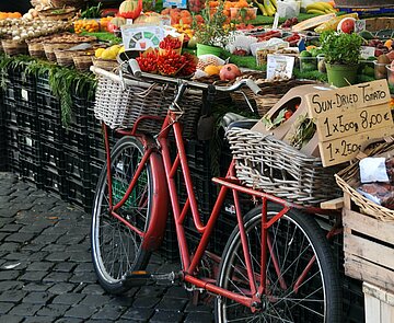 Ein Fahrrad lehnt an einen Marktstand. Die Fahrradkörbe (vorne und hinten) sind reich gefüllt mit Obst und Gemüse