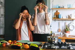 Zwei Menschen nebeneinander in einer Küche, die sich lächelnd Paprika-Stückchen vor die Augen halten