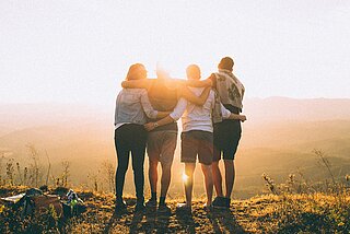 vier Menschen - mit dem Rücken zu sehen - vor einer Landschaft im Sonnenuntergang