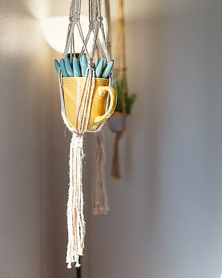 Eine mit Makramee-Schnüren an die Zimmerdecke gehangene gelbe Tasse, in der eine Pflanze (Sukkulente) wächst