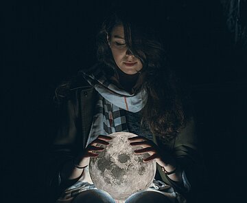 Weiblich gelesene Person in der Dunkelheit hält leuchtenden Globus in den Händen.