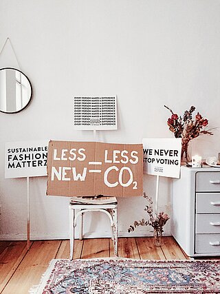 Demoplakate in einem Zimmer, auf einer zentral positionierten Pappe steht: Less New - Less CO2