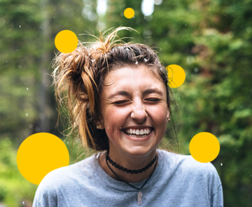 Portrait einer lachenden, weiblich gelesenen Person. Im Hintergrund sind Wald und gelbe Sphären.