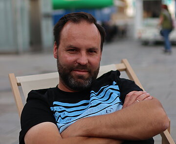 Porträt von Tim Preuß für die KA°-Community auf dem Karlsruher Marktplatz; junger Mann mit dunklen Haaren und Bart auf einem der KA°-Liegestühle sitzend, lächelnd