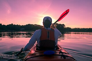Mann im Kayak, der auf einem See dem Sonnenuntergang entgegen paddelt
