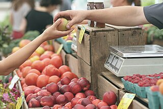 Marktstand, an dem jemand einen Apfel über den Tresen reicht, es sind nur die Hände, der Apfel und der Marktstand zu sehen