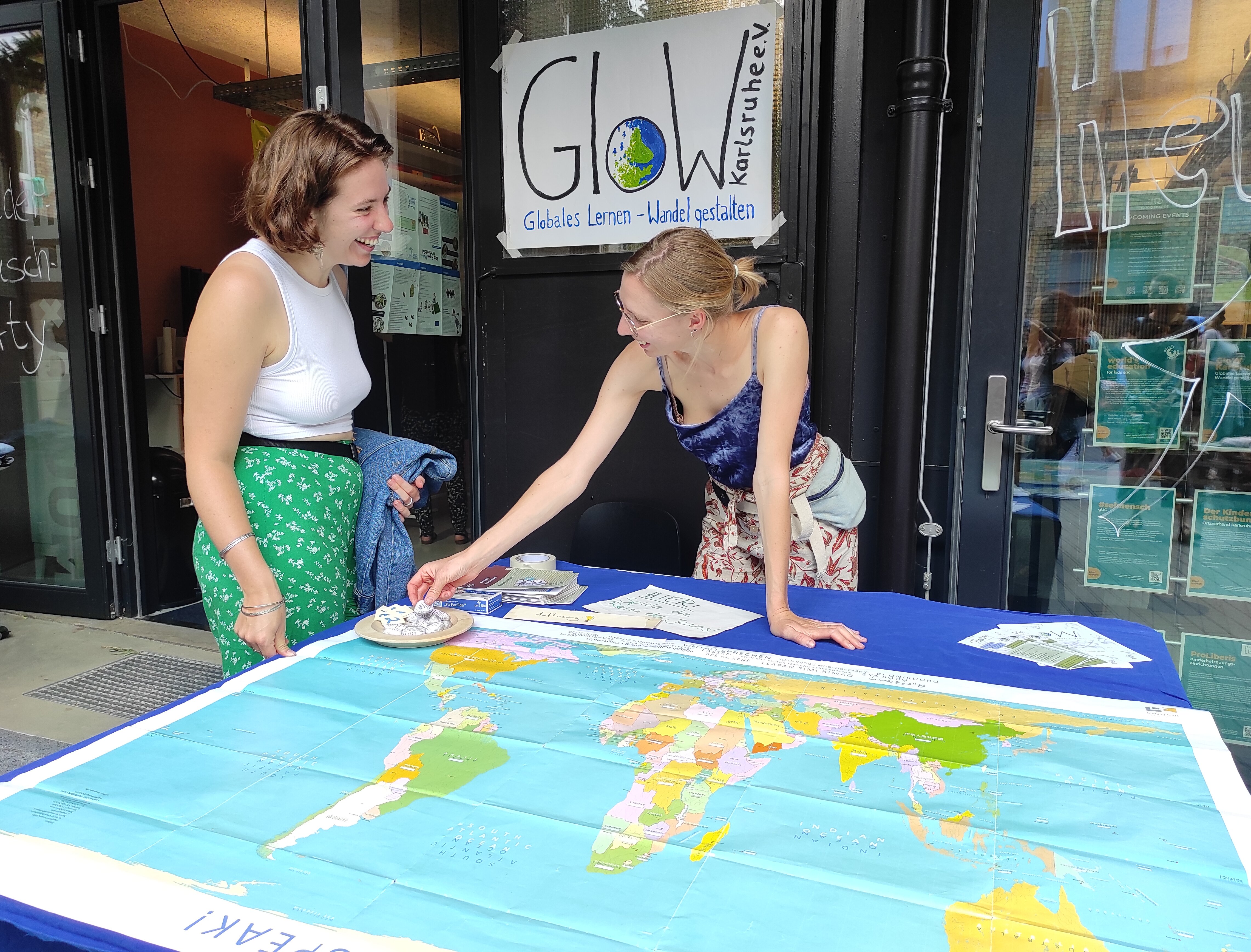 Zwei junge Frauen an einem Bildungsstand des GloW Karlsruhe e.V., vor ihnen eine große Weltkarte, im Hintergrund das GloW Karlsruhe e.V.-Logo hänend an einer Glastür