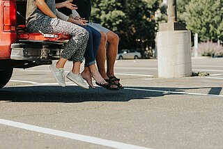 drei Personen, die auf einem Parkplatz Ladefläche eines Autos sitzen - man sieht nur die Beine hinunter baumeln